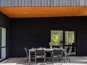 A modern outdoor dining area with a wooden deck, a table with chairs, and a large window offering a view of greenery. - Paradiset 2 by Interhome (Holmsjö)