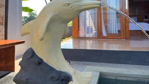 Una escultura de delfín con agua brotando de su boca, ubicada frente a una estructura de madera con puerta de vidrio y una piscina.