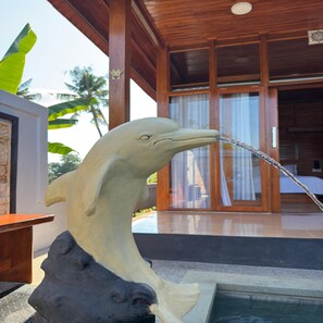 A dolphin sculpture with water spouting from its mouth, positioned in front of a wooden structure with a glass door and a pool. - Deepa Villa Lovina  (Buleleng)