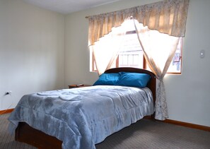 Une chambre à coucher avec un lit, une fenêtre avec des rideaux, une table de chevet et une prise électrique au mur.