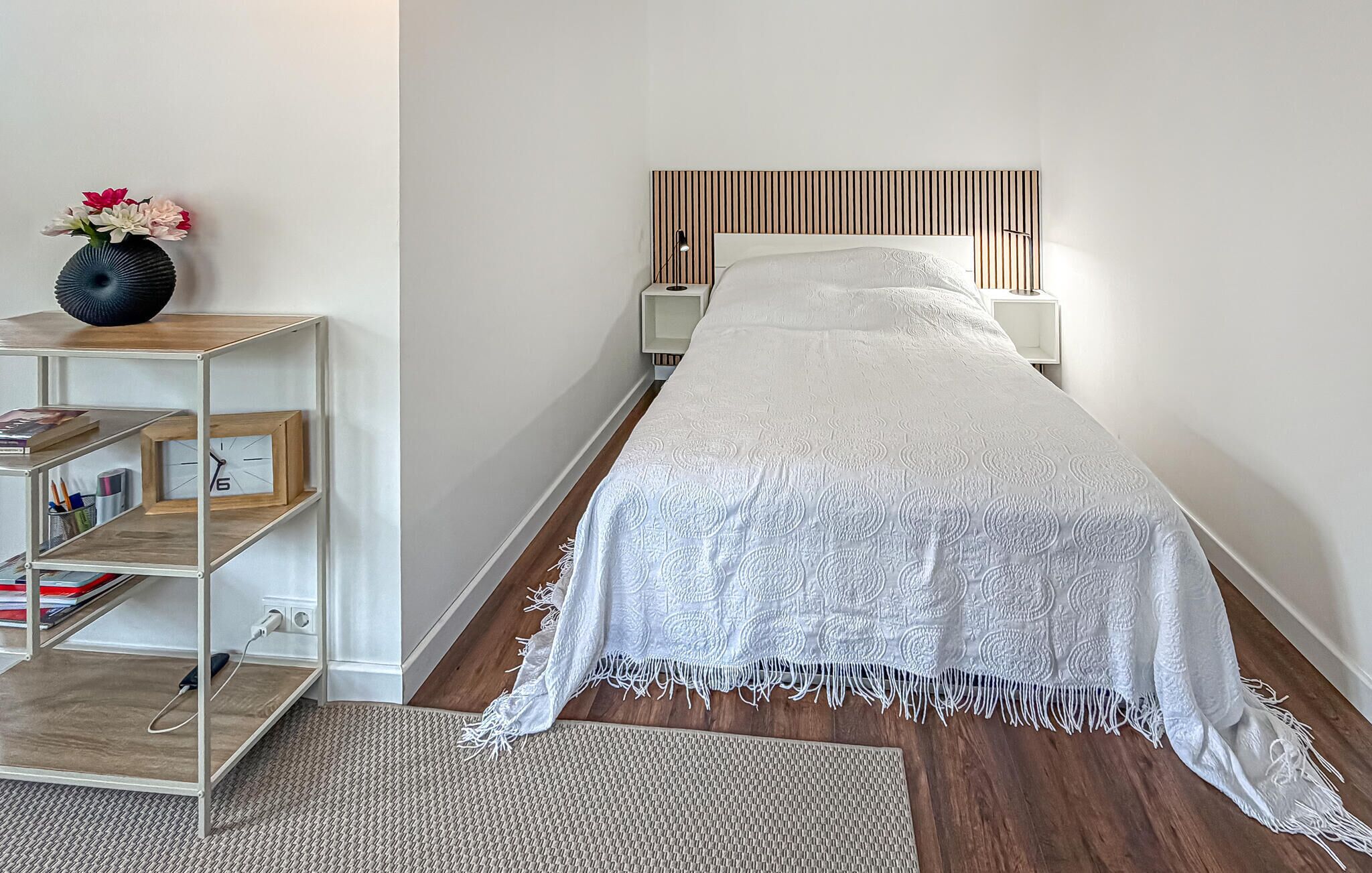 A bedroom with a bed, a bedside table, a lamp, a shelf with books, and a vase with flowers.