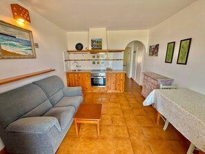A living room with a sofa, a coffee table, a kitchen area with cabinets and a stove, and a dining table with a tablecloth. - Suite with pool and sea views in Cala Morell (Cala'N Morell)