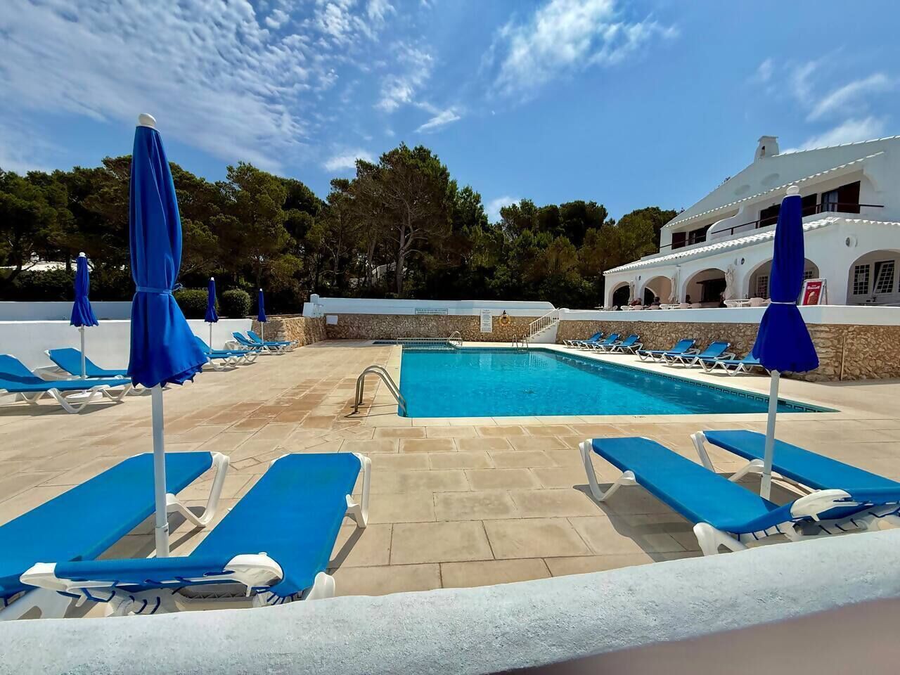 A pool area with blue lounge chairs and umbrellas, a stone-tiled deck, and a white building with arches in the background.