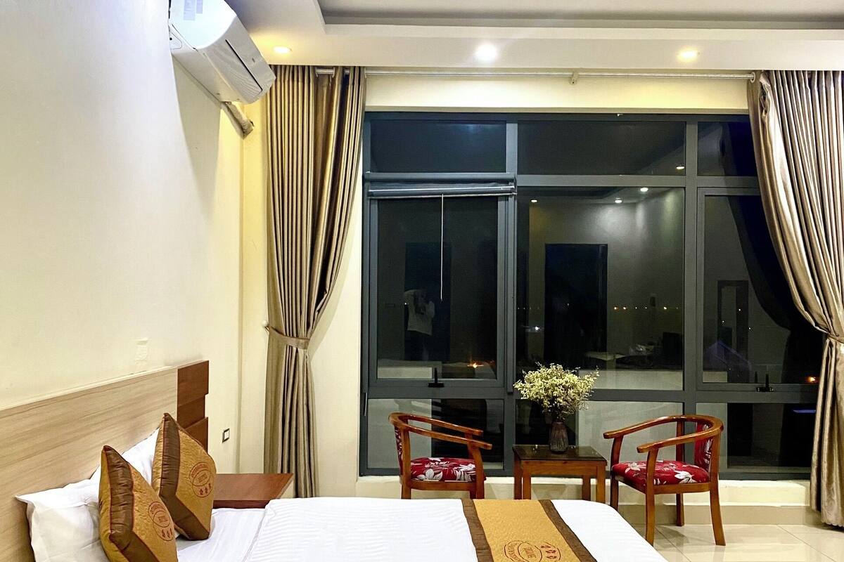 A hotel room with a large bed, two wooden chairs, a small table, and a view through large windows.