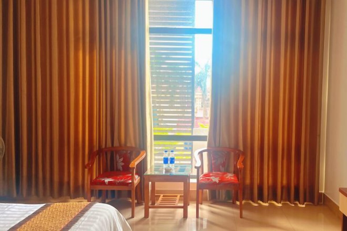 A hotel room with a bed, two chairs, a small table with water bottles, and large curtains.