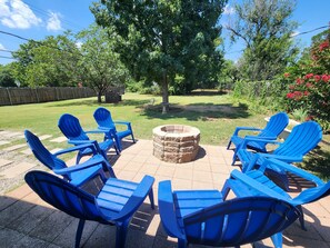 Eine Terrasse mit blauen Stühlen, die um eine Steinkaminfeuerstelle angeordnet sind, umgeben von einem Rasen und Bäumen.
