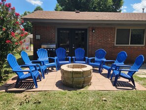 A patio with blue Adirondack chairs arranged around a stone fire pit, a grill, and a brick building with a window. - The Thunderdome Home (Norman)