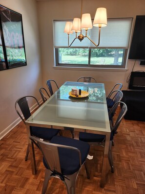 A dining area with a glass-top table, six chairs, a chandelier, and a window with blinds. - The Thunderdome Home (Norman)