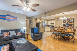 A living room with a ceiling fan, a dining area with wooden chairs, a kitchen with a refrigerator, and a basketball-themed wall decoration.