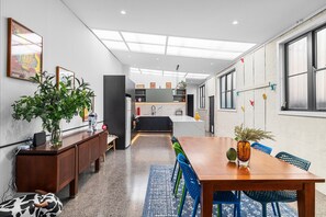 Eine moderne Küche mit einer zentralen Kochinsel, ein Essbereich mit einem Holztisch und blauen Stühlen sowie ein Wohnbereich mit einer Couch und einer Pflanze.