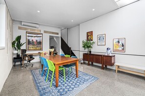 Ein moderner Büroarbeitsplatz mit einem Holztisch für Besprechungen, bunten Stühlen, einem blauen Teppich mit Muster und verschiedenen gerahmten Kunstwerken an den Wänden.