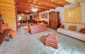 Ein gemütliches Wohnzimmer mit Holzbalkendecke, einer Couch, einem gemusterten Hocker und einem Esstisch mit Brot.