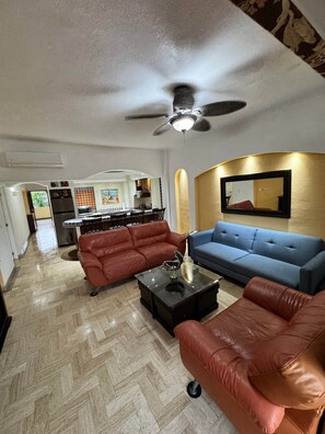 A living room with a ceiling fan, a blue sofa, red leather chairs, and a kitchen in the background. - Velas Vallarta Beach Front Resort (Puerto Vallarta)