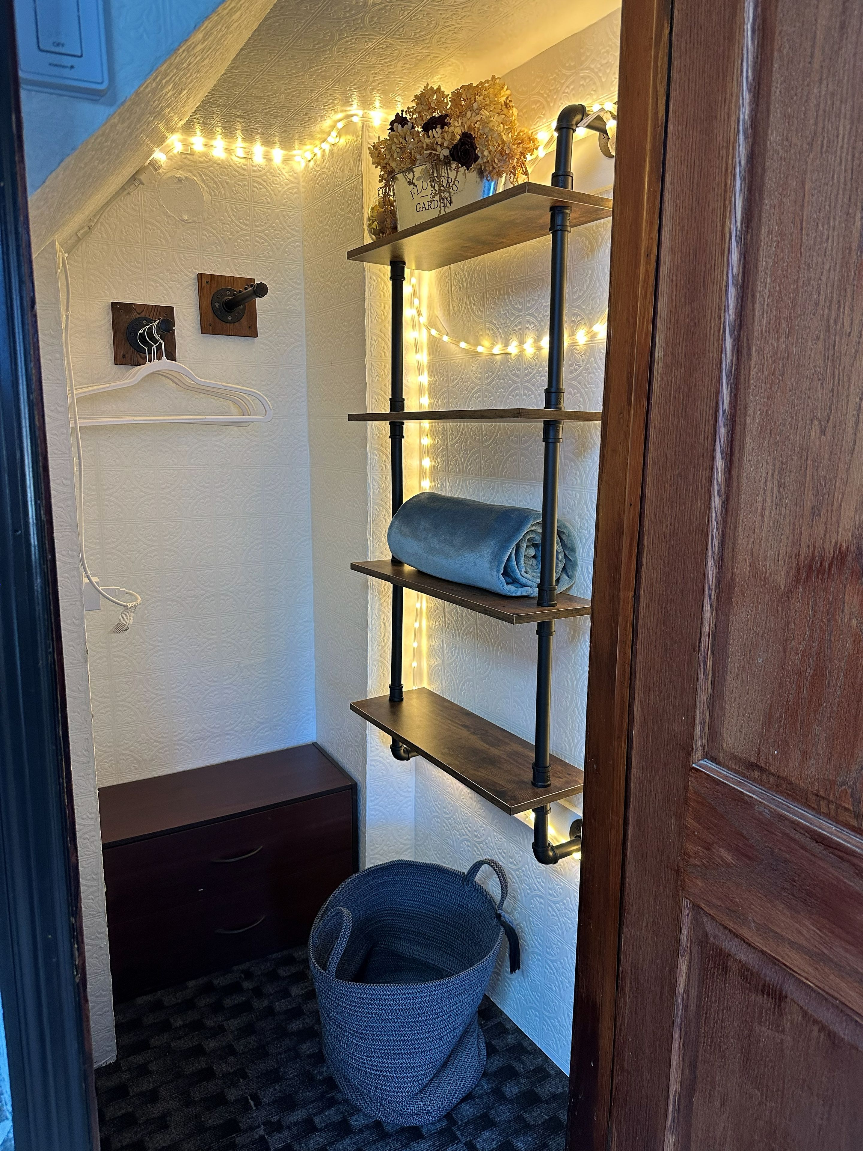 A corner of a room with a wooden bench, a wall-mounted shelf with a vase of flowers, and a basket.