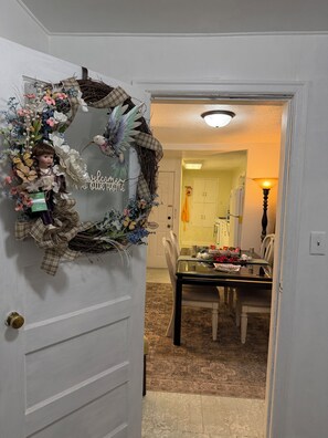 Un acceso con un adorno festivo, un comedor con mesa y sillas, y una cocina visible al fondo.