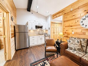 A compact kitchen with white cabinets, a stainless steel refrigerator, and a microwave. There's a wooden wall clock, a sofa with a patterned cushion, and a small table with a lamp.
