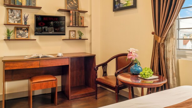 A room with a wooden desk, a chair, a small table with a vase and grapes, and shelves with books and framed pictures. - vina boutique hotel (Hue)