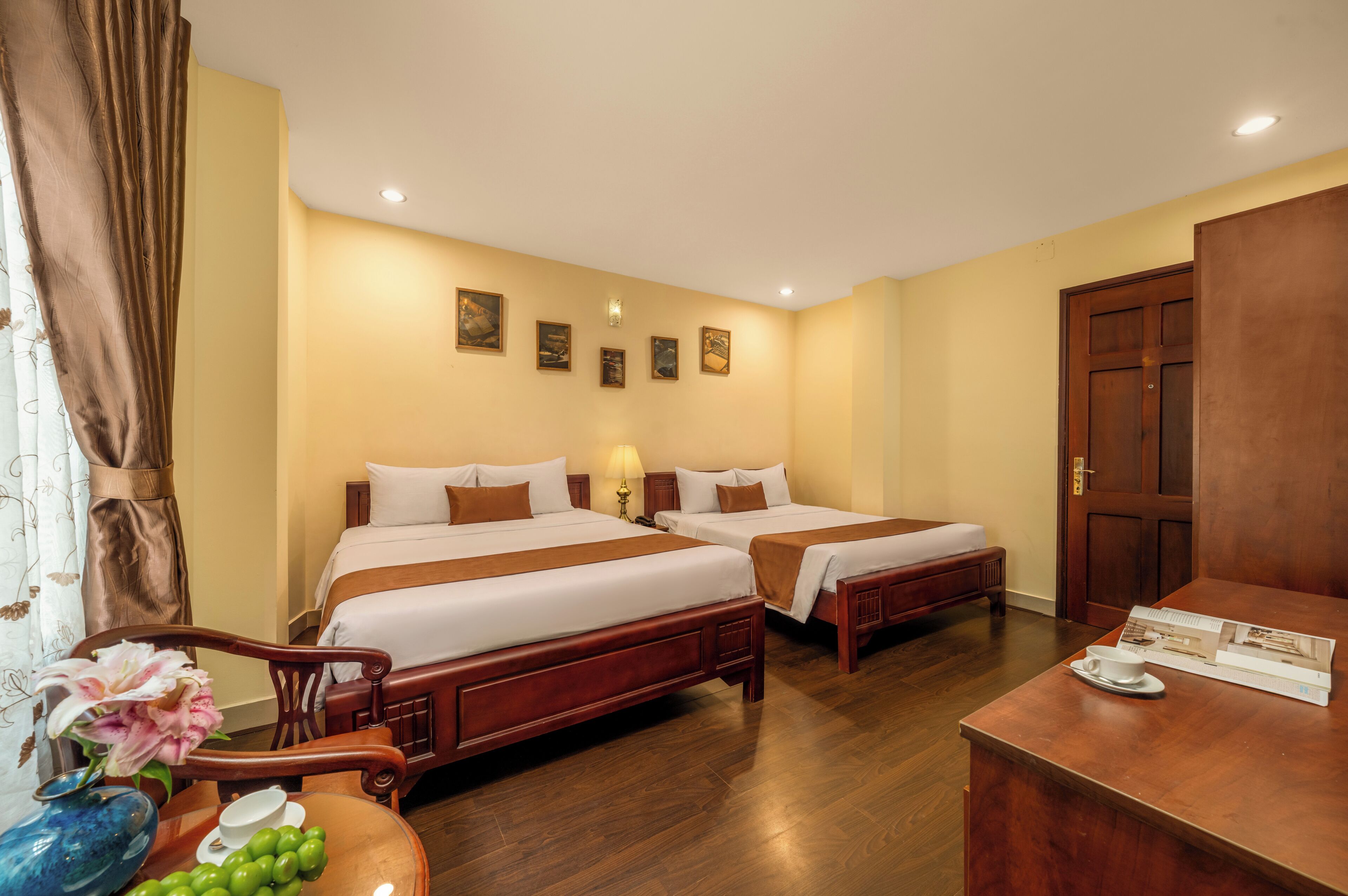 Een hotelkamer met twee bedden, een houten bureau, een stoel, een vaas met bloemen en een wand met ingelijste foto's.