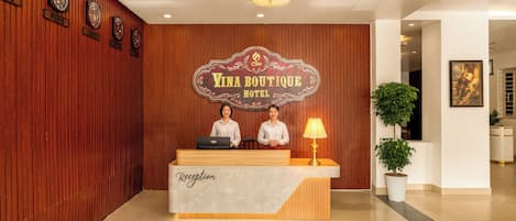 Una recepción con candelabro, paneles de madera, relojes y un cartel que dice 'Vina Boutique Hotel'.
