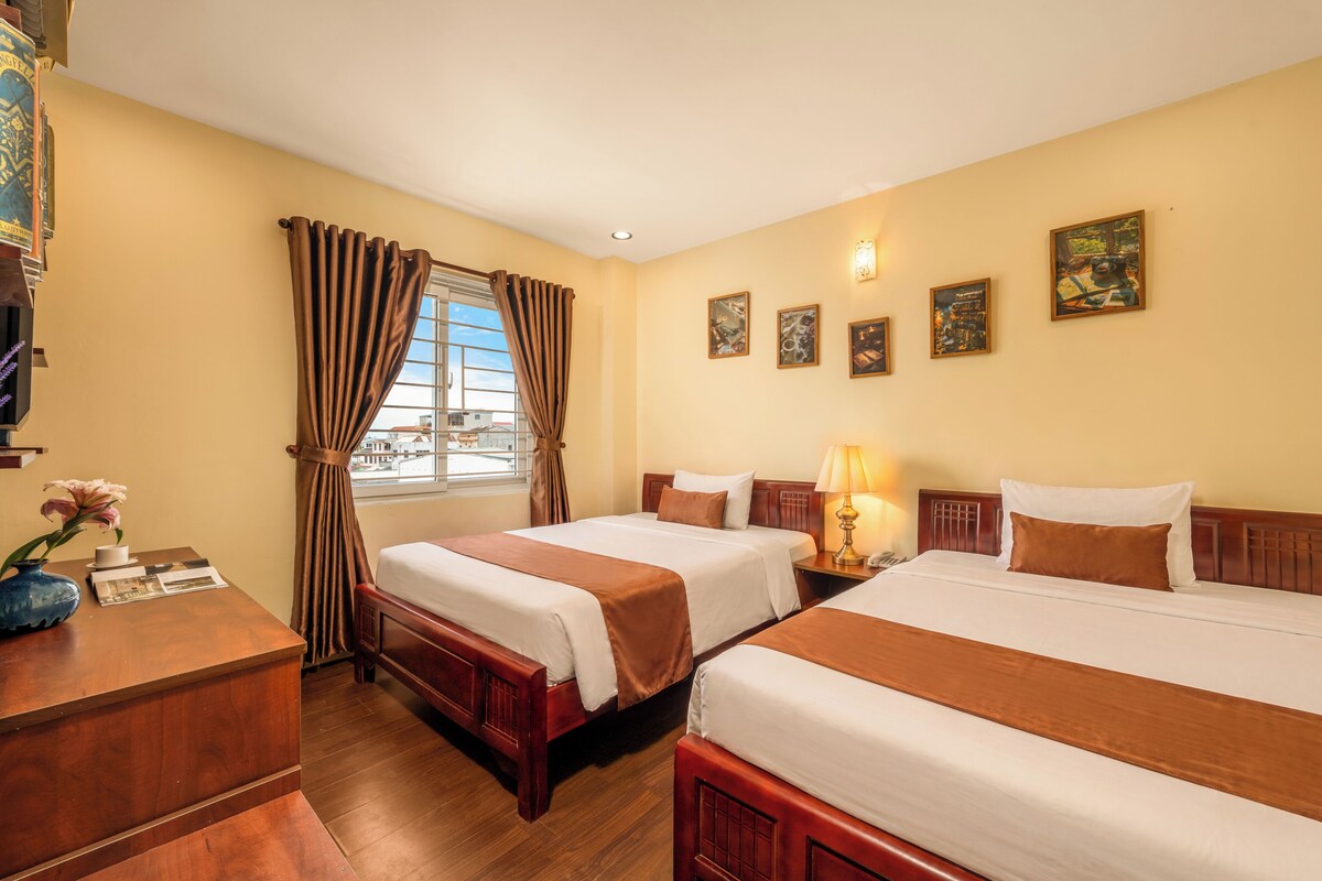 A hotel room with two beds, a desk, a window with curtains, and wall-mounted artwork.