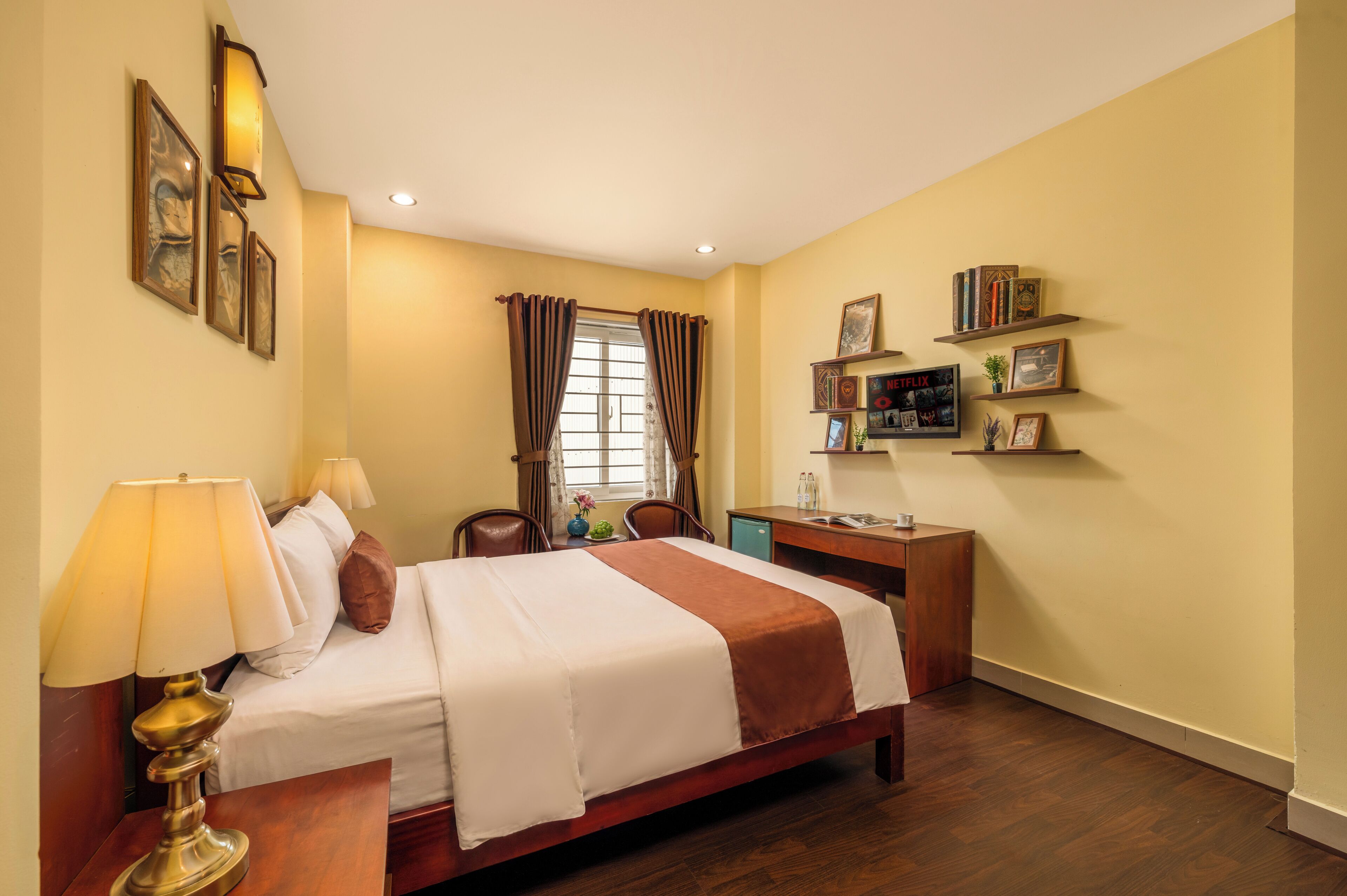 A hotel room with a bed, bedside lamp, desk, chair, and shelves with books and pictures.