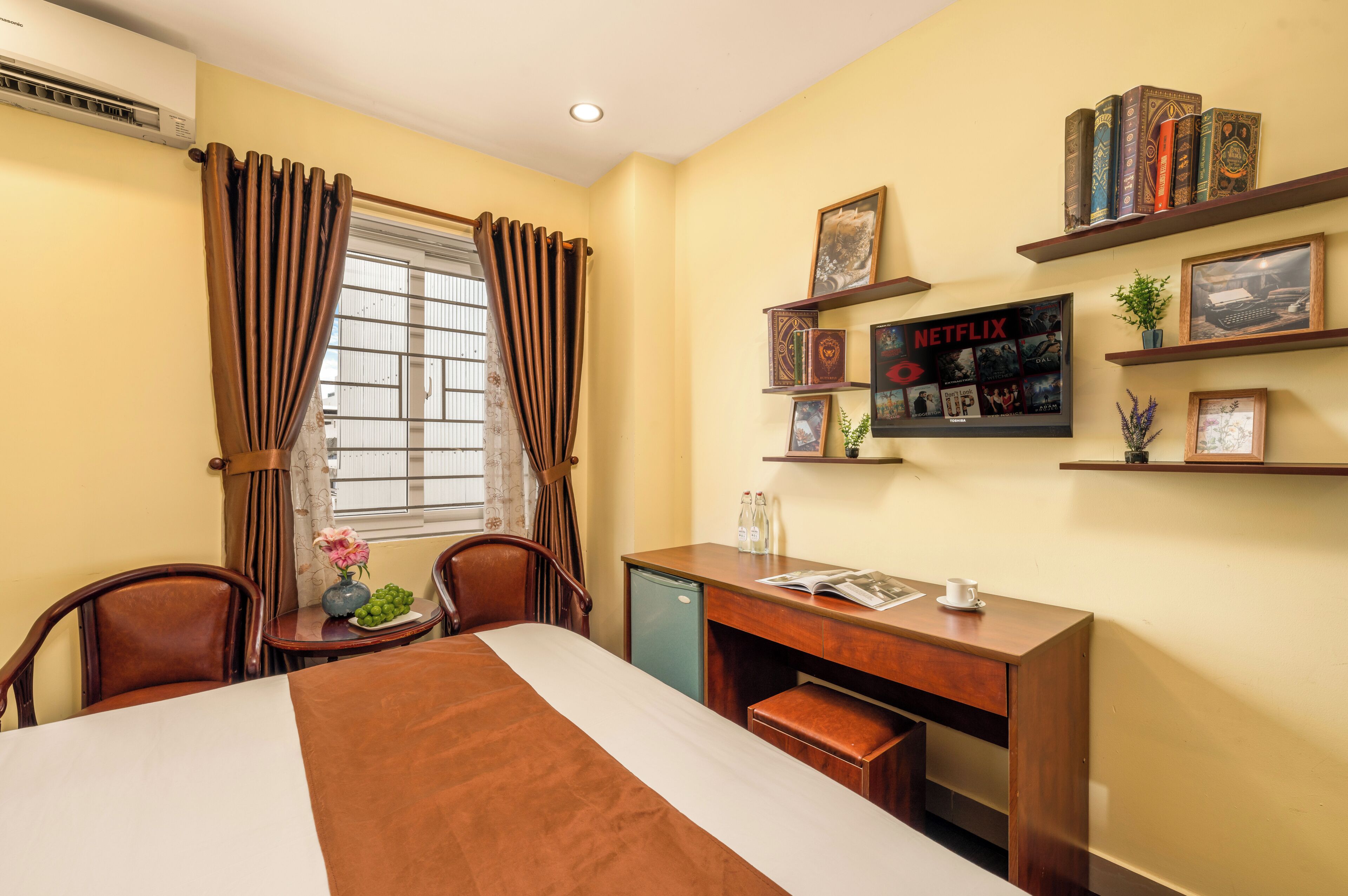 A hotel room with a bed, desk, chairs, a window with curtains, a wall-mounted shelf with books and pictures, and a wall-mounted air conditioner.