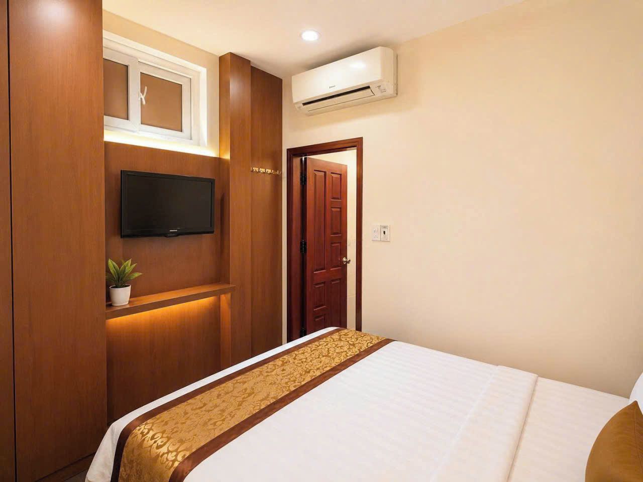 A hotel room with a bed, a TV mounted on the wall, a wooden wardrobe, a door, and an air conditioning unit.