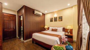 Habitación de hotel con una cama grande, dos mesitas de noche con lámparas, un armario de madera, un equipo de aire acondicionado y una pequeña mesa con un florero y fruta.