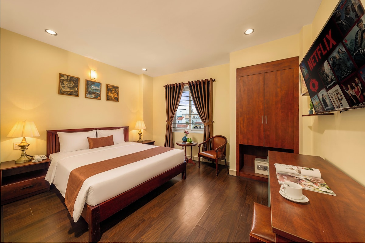 A hotel room with a bed, bedside lamps, a small table with a cup and magazine, a chair, a wardrobe, and a window with curtains.