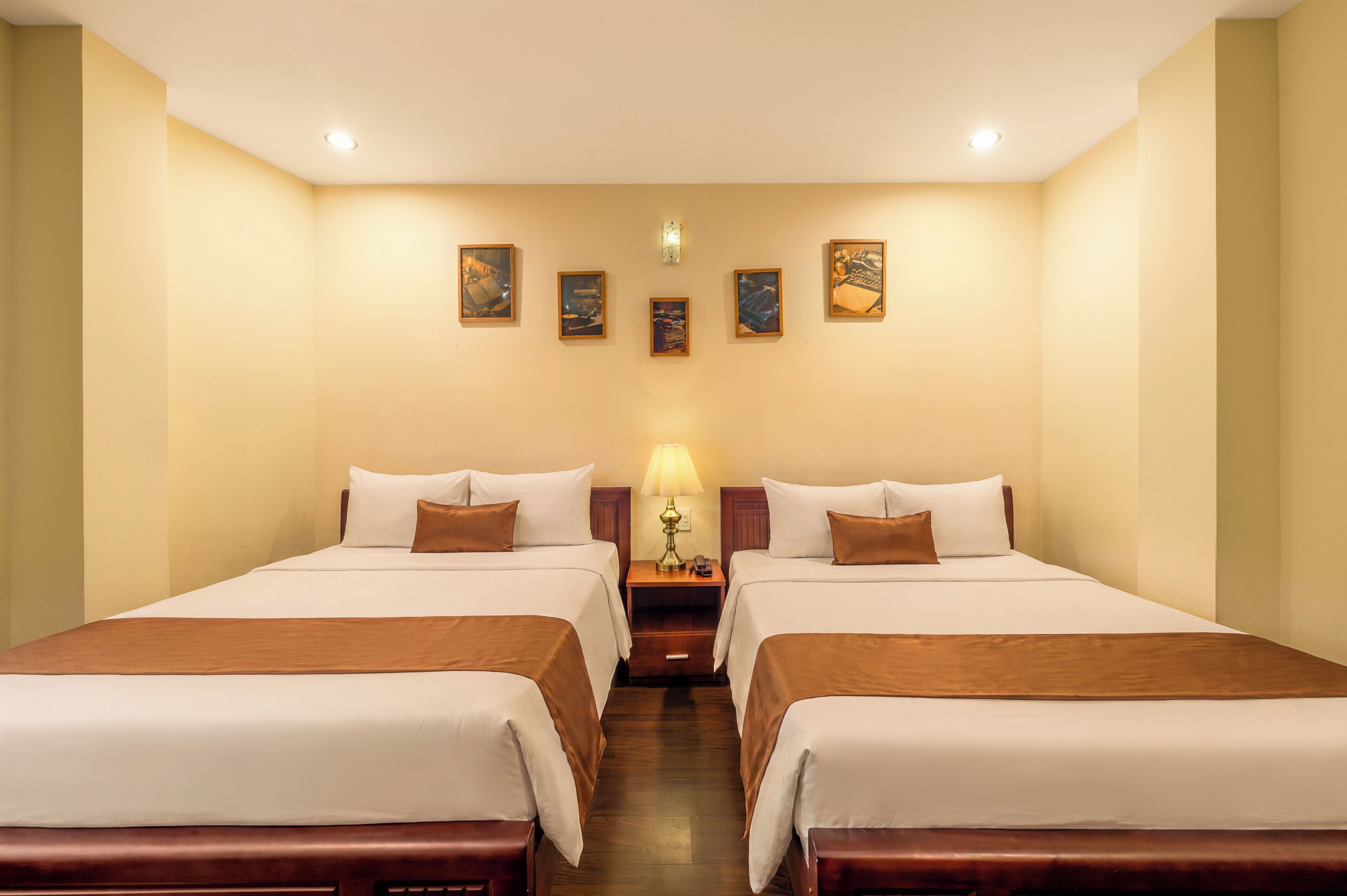Habitación de hotel con dos camas, una mesita de noche, una lámpara y cuatro cuadros en la pared.