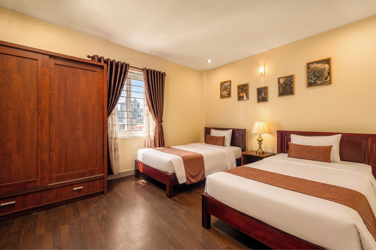 A hotel room with two beds, wooden wardrobe, curtains, wall-mounted lights, and framed pictures.
