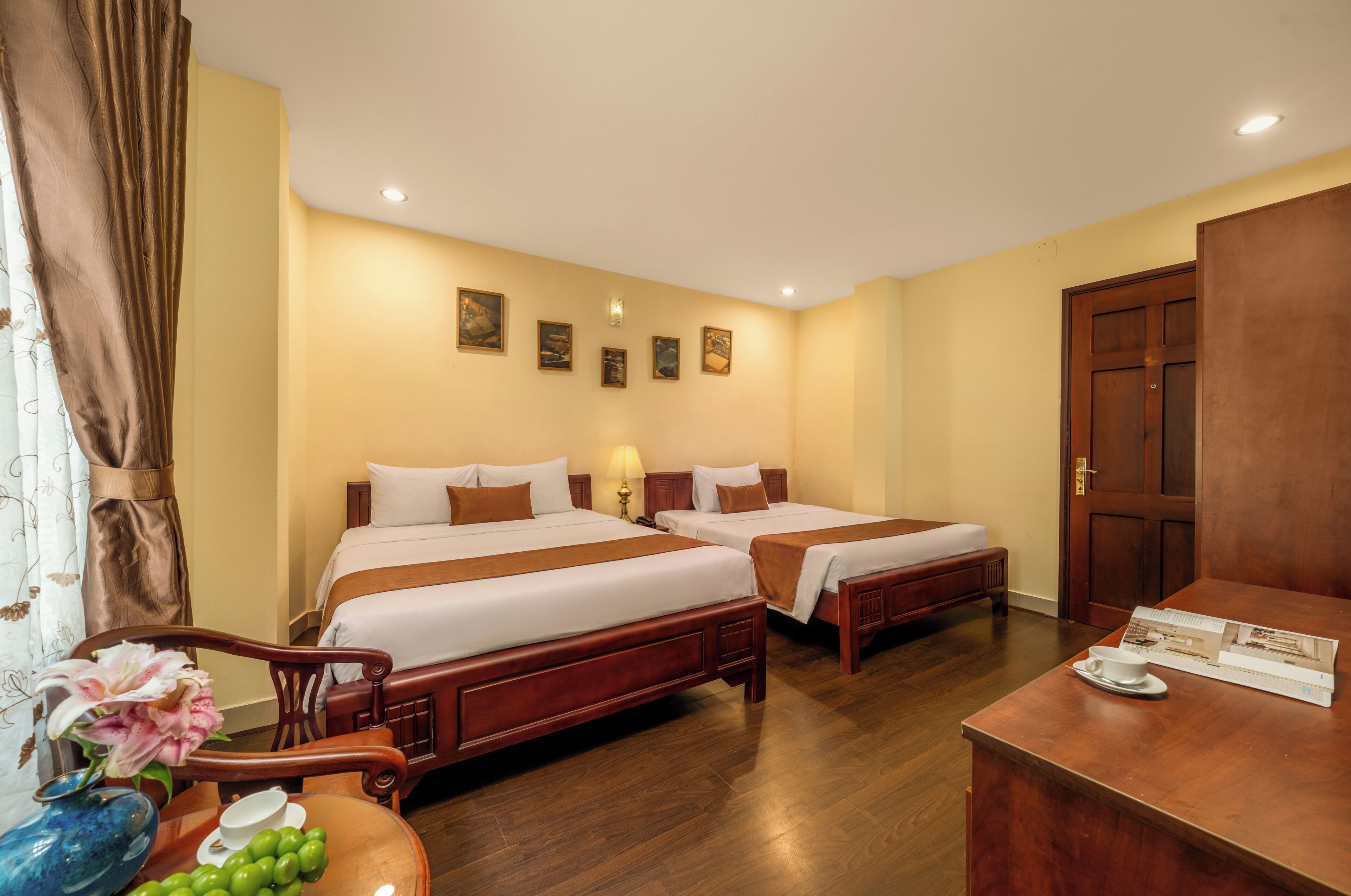A hotel room with two beds, a wooden desk, a vase with flowers, and a framed picture on the wall.