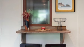 Un coin repas avec une table en bois, deux chaises noires, un vase contenant des fleurs séchées, un bol rouge et un tableau encadré accroché au mur.