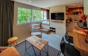 A compact room with a bed, a TV, a small kitchen area, and a window with a view of greenery. - Studio proche de la mer (Saint-Quay-Portrieux)