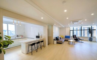 Cocina moderna con una isla central y taburetes, un espacio de estar con sofá y sillones, y ventanas amplias con vistas a la ciudad.