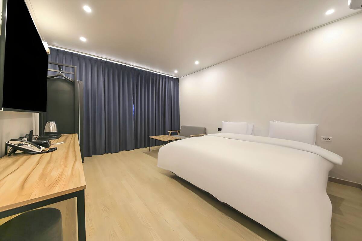 A modern hotel room with a wooden desk, a bed, a chair, and a TV mounted on the wall.