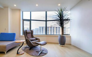 Un salon moderne avec une grande fenêtre, un fauteuil gris, un canapé bleu et une grande plante en pot.