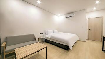 A modern hotel room with a bed, a grey sofa, a small table, and an air conditioning unit.