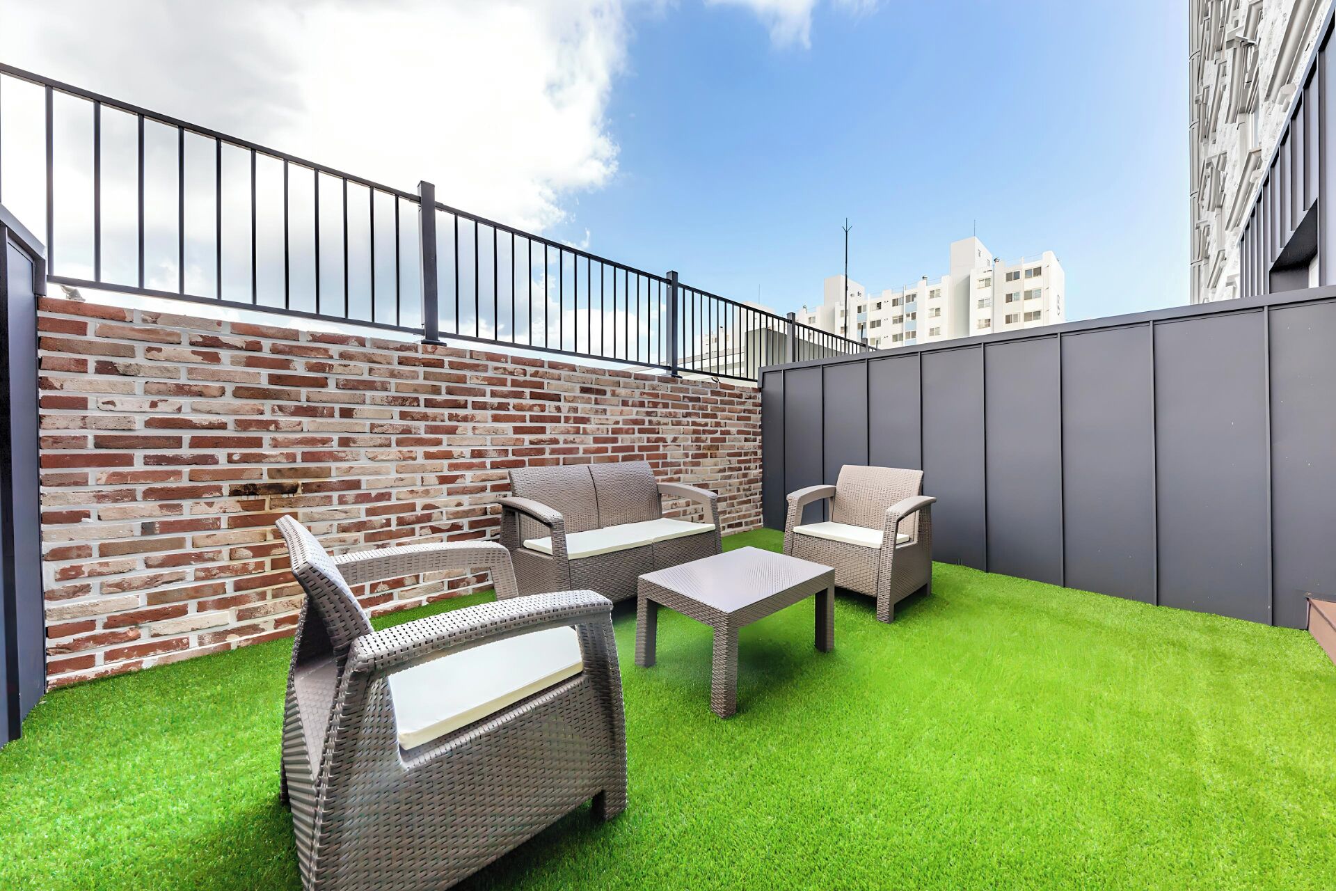 Un patio en la azotea con mobiliario de mimbre, césped artificial, pared de ladrillo y baranda de metal.