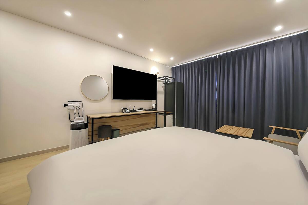 A modern hotel room with a large bed, a desk with a chair, a flat-screen TV, a round mirror, and a window with curtains.
