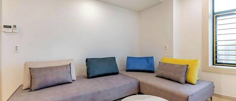 Un salon moderne avec un canapé gris, un pouf blanc et une fenêtre équipée de stores.