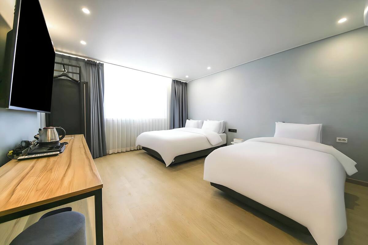 A modern hotel room with two beds, a desk with a kettle, a flat-screen TV, and a window with curtains.