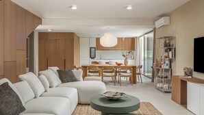Uma sala de estar moderna com um sofá seccional, uma área de jantar com mesa e cadeiras de madeira, e uma cozinha ao fundo.