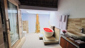 A modern bathroom with a glass shower enclosure, a freestanding bathtub with red flowers, a wooden vanity, and a skylight. - Bonito Suite Villa (Ubud)