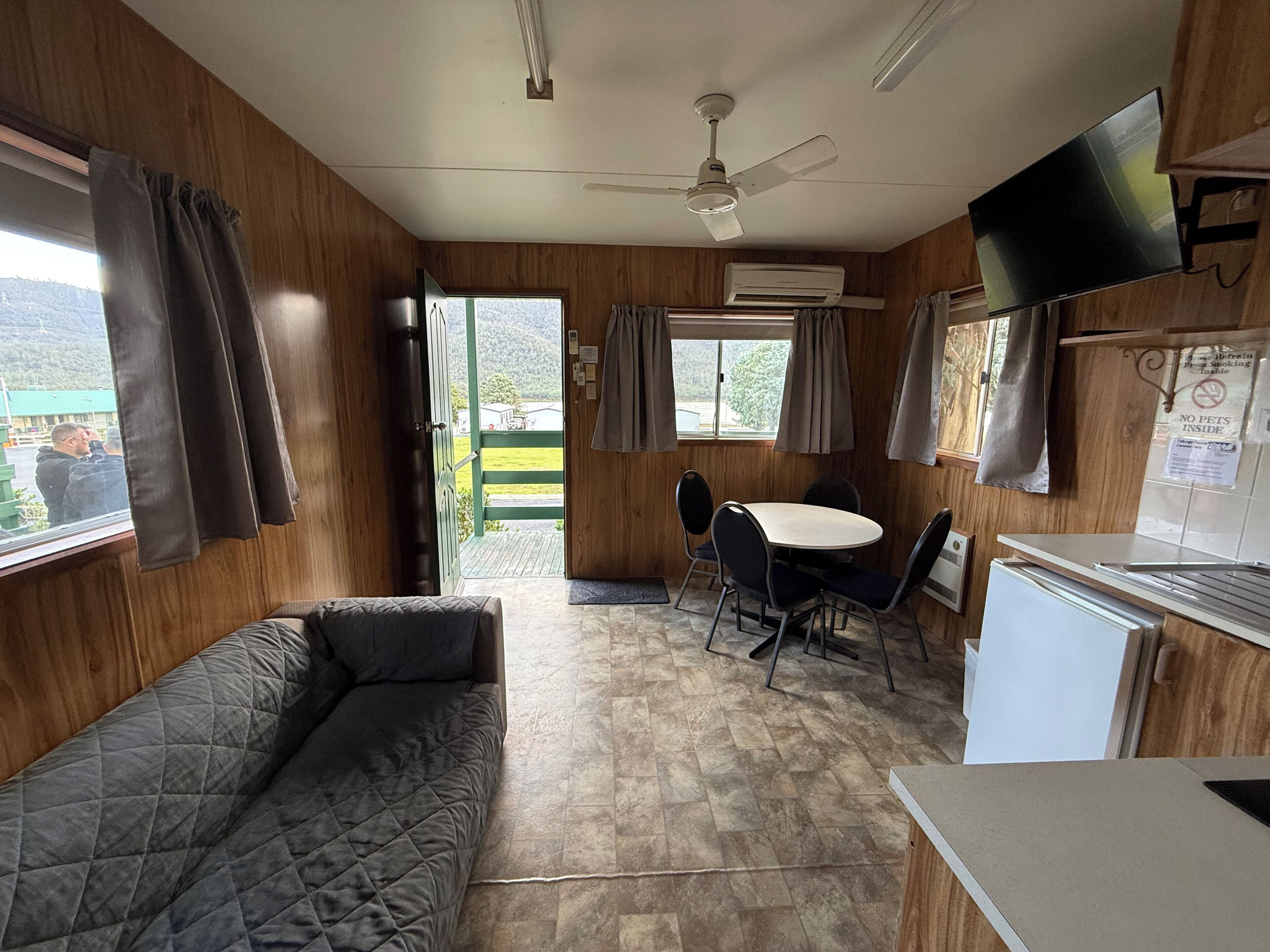 A room with a sofa, a table with chairs, a ceiling fan, an air conditioning unit, a television, and a window with curtains.