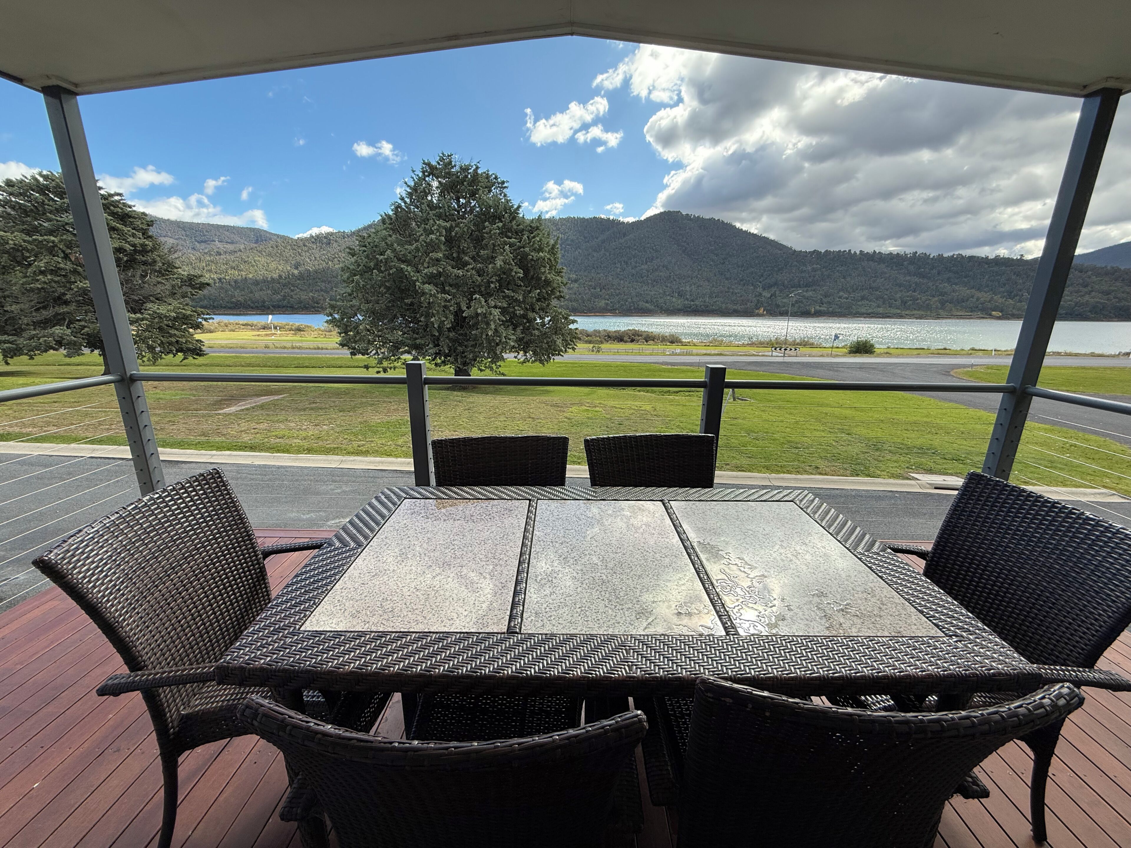 A covered outdoor seating area with a table and chairs, overlooking a lake and mountains.