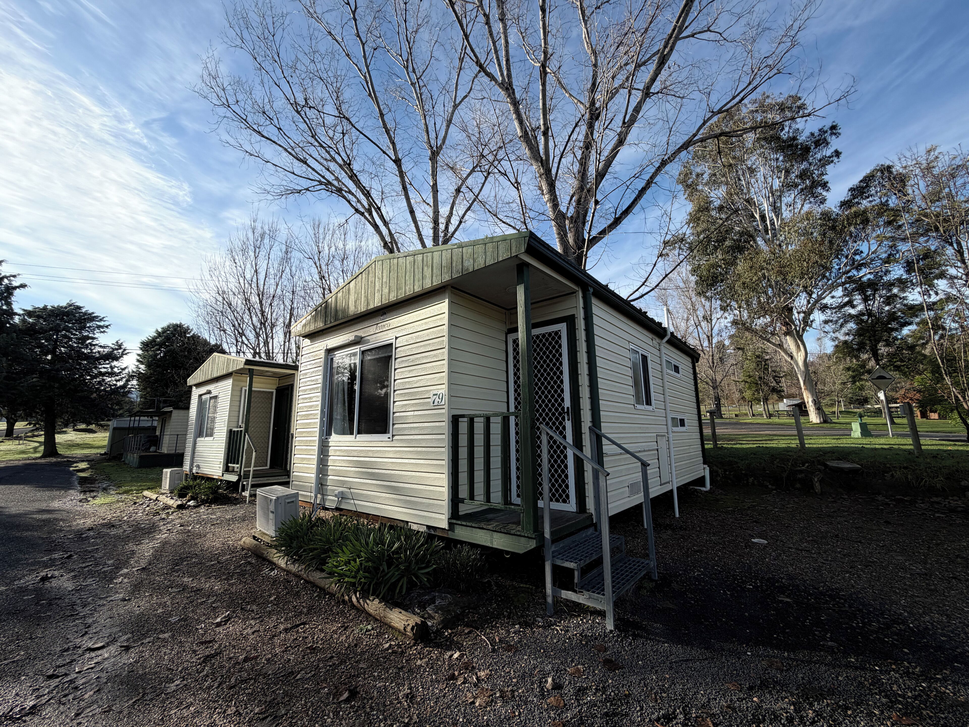 A small, single-story mobile home with a porch and a fenced yard, surrounded by trees.