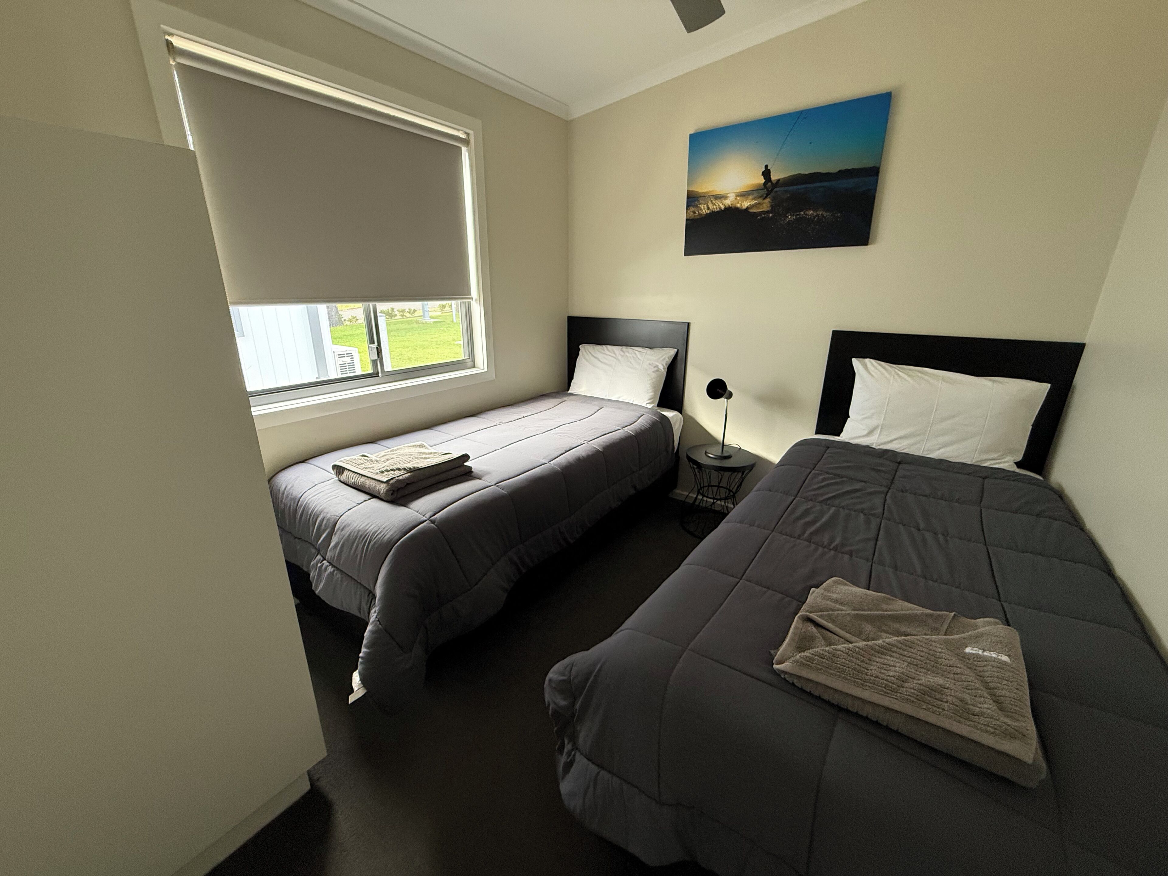 Ein Schlafzimmer mit zwei Betten, einem Fenster mit Jalousien, einem Nachttisch mit Lampe und einem Bild an der Wand.