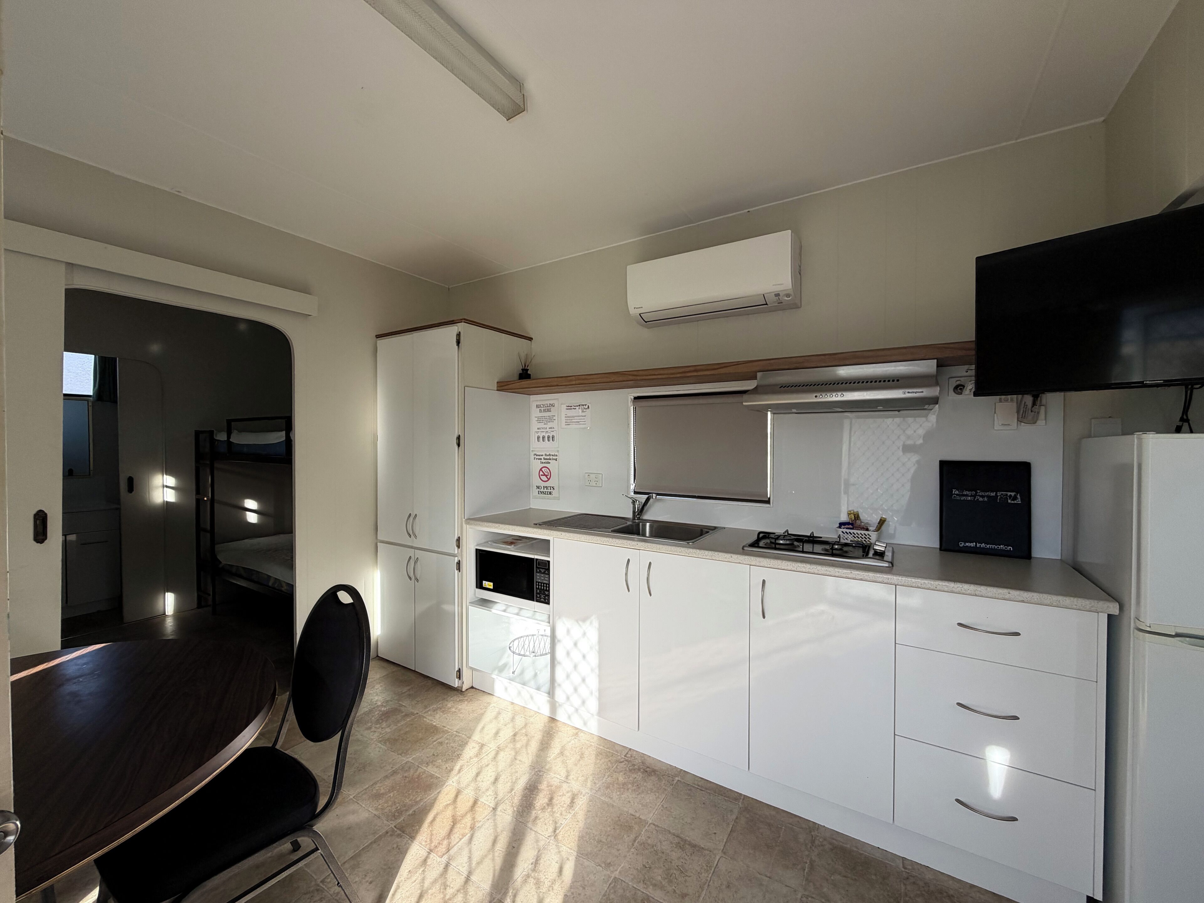 A compact kitchen with white cabinets, a gas stove, a microwave, and a sink. There is a dining table with chairs and a TV mounted on the wall.