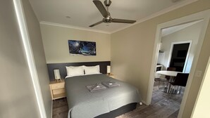 A bedroom with a bed, bedside tables, a ceiling fan, a painting on the wall, and a view into the dining area. - Tiny Away Escape at Talbingo Alpine (Talbingo)